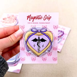 Può includere: Un'impugnatura magnetica a forma di cuore per e-reader, telefoni e tablet su un cartoncino rosa con motivo a fiocchi. L'impugnatura presenta un cuore viola chiaro con bordo giallo, un fiocco viola e l'illustrazione nera di una spada alata.