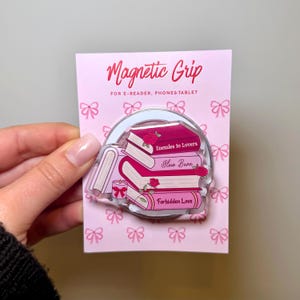 Op de afbeelding: Een roze magnetische grip voor e-readers, telefoons en tablets. De grip toont een stapel boeken met de titels "Enemies to Lovers", "Slow Burn" en "Forbidden Love". Het ontwerp bevat rozen en een kaars.