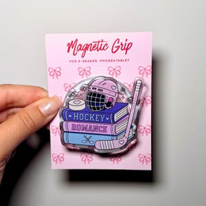 Könnte beinhalten: Ein runder magnetischer Griff auf einer rosa Karte mit dem Text Magnetic Grip FOR E-READER, PHONE & TABLET. Der Griff zeigt einen Stapel aus drei Büchern mit der Aufschrift HOCKEY ROMANCE, einen rosa Eishockeyhelm, einen Puck und einen Schläger.