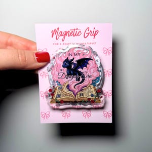 May include: A pink magnetic grip for e-readers or tablets. The grip features a black dragon with purple wings, the text "IN MY Dragon Era", and an open book with roses. The words "Magnetic Grip" are at the top.