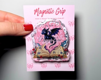 May include: A clear acrylic magnetic grip for e-readers and tablets on a pink backing card with a bow pattern. The grip features a black dragon with purple wings emerging from an open book, surrounded by pink smoke and the text In My Dragon Era.
