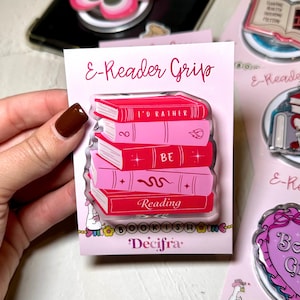 Könnte beinhalten: Ein pinkfarbener und roter E-Reader-Griff mit einem Stapel Bücher und dem Schriftzug "I'd Rather Be Reading". Der Griff hat ein klares, glänzendes Finish und ist auf einer pinkfarbenen Rückkarte mit dem Text "E-Reader Grip" und "Decifra".