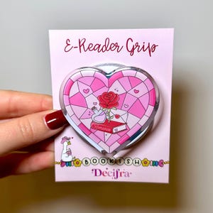 May include: A heart-shaped E-Reader Grip on a pink display card. The grip features a stained-glass design with a red rose, a potion bottle, and a red book labeled Romance. The card displays the text E-Reader Grip, BOOKISH, and Decifra.