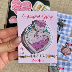 Può includere: Un supporto E-Reader Grip su un cartoncino rosa chiaro con l'illustrazione di una boccetta a forma di cuore. La boccetta è viola con le scritte Romance, Book Essence e True Love. In basso a sinistra c'è una piccola oca bianca con cappello rosa.