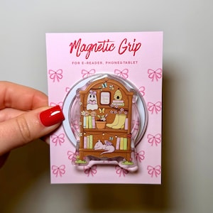 Op de afbeelding: Een roze kaart met een magnetische grip voor e-readers, telefoons en tablets. De grip heeft een grillige illustratie van een boekenplank met boeken, een spook, een draak en een bijenkorf. De tekst "Magnetic Grip" staat bovenaan.