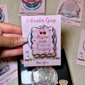 Puede incluir: Un soporte rosa para lector de libros electrónicos con el texto "Buy me books & tell me I'm pretty" e ilustraciones de cerezas. El soporte está en una tarjeta rosa con el texto "E-Reader Grip" y la marca "Decifra". Otros soportes similares son visibles en el fondo.