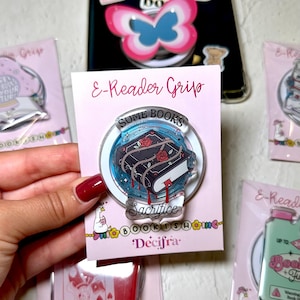 Può includere: Un supporto circolare in acrilico per e-reader su un cartoncino rosa chiaro con l'etichetta E-Reader Grip. Il supporto presenta l'illustrazione di un libro scuro con rose rosse, spine e gocce rosse, con il testo SOME BOOKS Sacrifice stampato.
