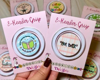 May include: Two circular e-reader grips on pink backing cards. One grip features a light blue plaid background with a green book and the text Anti Social Book Club. The other grip has a peach plaid background with the text Plot twist in black.