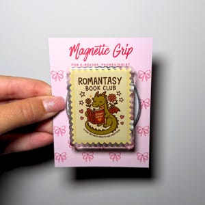 Op de afbeelding: Een rechthoekige magnetische grip voor e-readers, telefoons en tablets op een roze kaart met strikjespatroon. De grip heeft een postzegelvormig ontwerp met een groene draak die een boek leest, de tekst ROMANTASY BOOK CLUB en bloemen.