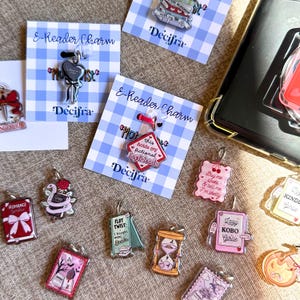 Op de afbeelding: Een verzameling acryl e-reader bedels met diverse ontwerpen, waaronder een hartvormige lolly, een ruitvormig bordje met de tekst this holds my fictional boyfriends, boeken, een zandloper en postzegels met teksten zoals Romance Girlie.