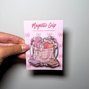 Può includere: Un supporto magnetico rosa per e-reader, telefoni e tablet su un cartoncino. Il supporto presenta un'illustrazione in acrilico trasparente di una borsa a quadretti rosa piena di libri, un fiocco, piccoli fiori e una tazza.