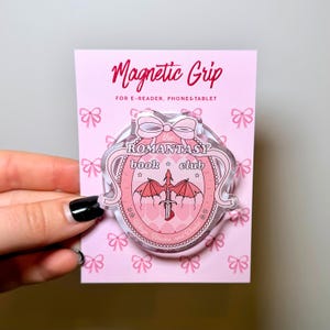 Può includere: Un supporto magnetico circolare in acrilico su un cartoncino rosa con un motivo a fiocchi. Il supporto presenta una cornice ovale rosa con un fiocco in alto, un grafico con spada e ali di drago e il testo "the ROMANTASY book club".