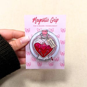 Può includere: Un supporto magnetico rosa e trasparente per e-reader, telefoni e tablet. L'impugnatura presenta un design a forma di bottiglia a cuore riempita di liquido rosso e fiamme, con le parole "slow burn essence" su un'etichetta. La confezione è rosa con illustrazioni di fiocchi.