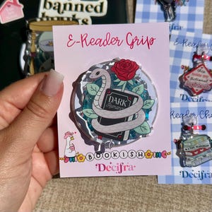 May include: A circular e-reader grip on a light pink backing card labeled E-Reader Grip. The grip features a white snake coiled around a black book titled Dark Romance, topped with a red rose and green leaves, set against a blue glitter background.