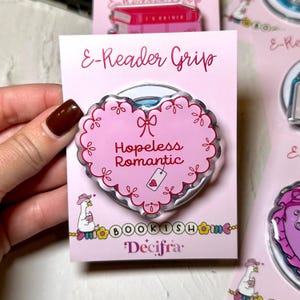 Może przedstawiać: Różowy uchwyt do czytnika e-booków w kształcie serca z napisem "Hopeless Romantic" i małą zawieszką z sercem. Uchwyt ma falisty brzeg i wzór kokardy. Karta zawiera również tekst "BOOKISH" i "Décifra".