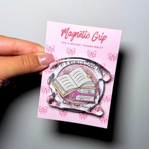 Könnte beinhalten: Ein transparenter magnetischer Griff aus Acryl für E-Reader, Handys und Tablets auf einer rosa Karte mit Schleifenmuster. Der Griff zeigt eine Illustration von gestapelten Büchern und den Text: I'M IN A REALLY GOOD PLACE in my book, not mentally.