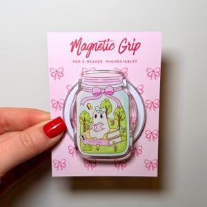 Könnte beinhalten: Ein rosa Magnetgriff für E-Reader, Telefone und Tablets. Der Griff zeigt ein Glasdesign mit einem Geist, der in einer Frühlingsszene ein Buch liest. Der Text "Magnetic Grip" und "Spring" sind sichtbar.