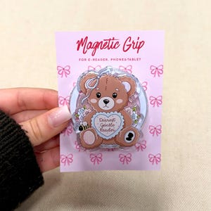 Puede incluir: Un soporte magnético rosa para lectores electrónicos, teléfonos y tabletas. Presenta un diseño de oso de peluche marrón con lazo, flores y el texto "Dearest Gentle Reader". El embalaje es rosa con lazos.