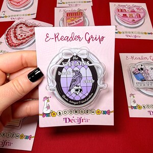 May include: An acrylic E-Reader Grip featuring a circular design with a white bow, a purple stained-glass window pattern, a black raven, and a skull. The text reads: the VILLAINS book club, Books, betrayal & black feathers. It is on a display card.