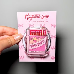 Op de afbeelding: Een roze magnetische grip voor e-readers, telefoons en tablets met een luciferdoosje-illustratie, de tekst Slow Burn en made for readers. De achterkaart is lichtroze met een strikjespatroon en de tekst Magnetic Grip.