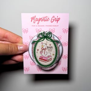 May include: A circular magnetic grip for e-readers, phones, and tablets on a pink backing card with a bow pattern. The grip features a white duck illustration, a stack of books, and the text "BOOK TROVERT" inside a dark green scalloped frame.
