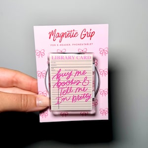 Op de afbeelding: Een rechthoekige magnetische grip voor e-readers, telefoons en tablets op een roze kaart met strikjespatroon. De grip heeft een bibliotheekkaartontwerp met de tekst "buy me books & Tell me I'm pretty" in felroze handgeschreven letters.