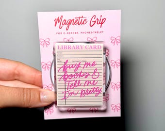 May include: A rectangular magnetic grip for e-readers, phones, and tablets on a pink backing card with a bow pattern. The grip features a vintage library card design with the text "buy me books & Tell me I'm pretty" written in bright pink script.