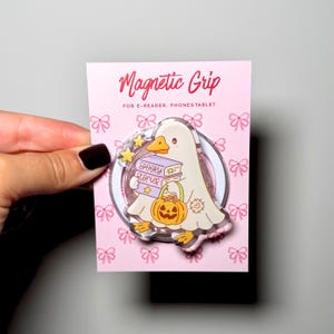 Può includere: Un supporto magnetico circolare per e-reader, telefoni e tablet su un cartoncino rosa con motivo a fiocchi. Il supporto raffigura un'anatra vestita da fantasma che tiene in mano libri con la scritta Spooky Stories e un secchiello a zucca.