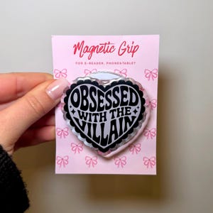 May include: A pink card with a magnetic grip for e-readers, phones, and tablets. The grip is heart-shaped with the words "OBSESSED WITH THE VILLAIN" in black and white. The card has a pink background with small pink bows.