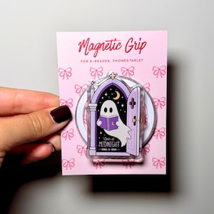 Può includere: Un supporto magnetico per e-reader, telefoni e tablet su una carta rosa con motivo a fiocchi. Il supporto raffigura un fantasma che legge un libro viola all'interno di un arco gotico con il testo Open at MIDNIGHT, su un cielo stellato.