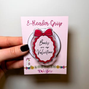 Può includere: Un accessorio E-Reader Grip su un cartoncino espositivo rosa chiaro. L'impugnatura presenta una forma ovale smerlata con un fiocco rosso in alto e il testo Books are my Valentine. Il cartoncino include una piccola illustrazione di un'oca e il marchio Decifra.