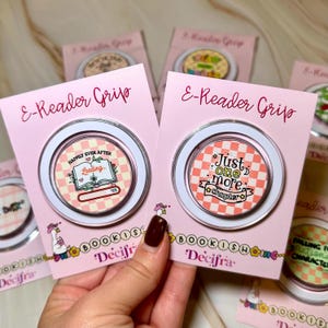 Peut inclure: Supports de liseuse électroniques roses avec des bordures blanches et des motifs décoratifs. L'un des supports présente un livre avec le texte "Happily Ever After" et "Loading". L'autre support porte le texte "Just one more chapter" sur un fond à carreaux.
