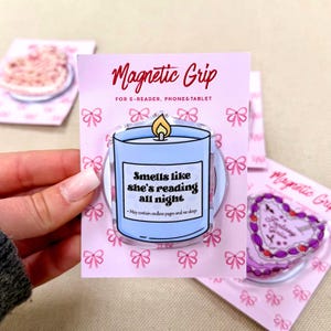 May include: A light blue candle-shaped magnetic grip on a pink backing card with a repeating bow pattern. The candle features a yellow flame and text that reads: Smells like she's reading all night. May contain endless pages and no sleep.