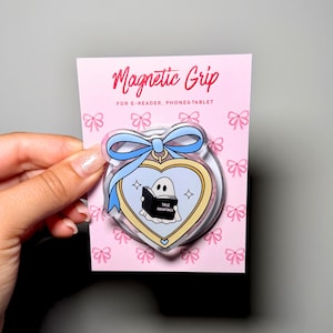 Könnte beinhalten: Ein runder magnetischer Griff für E-Reader, Handys und Tablets auf einer rosa Karte mit Schleifenmuster. Der Griff hat einen herzförmigen Rahmen mit blauer Schleife und zeigt eine Illustration eines Geistes, der ein Buch namens True Hauntings liest.