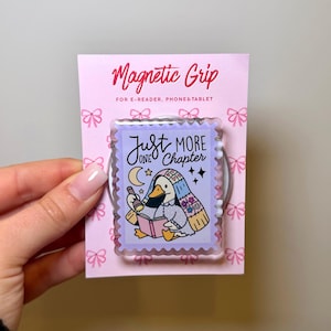 Op de afbeelding: Een roze magnetische grip voor e-readers, telefoons en tablets. De grip heeft een cartoon eend die een boek leest met de tekst "Just One More Chapter". Het ontwerp bevat een maan, sterren en bloemmotieven.