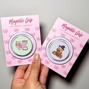 Può includere: Due accessori circolari Magnetic Grip per e-reader, telefoni e tablet su cartoncini rosa con motivi a fiocco bianchi. Uno presenta un libro rosa e una clessidra con il testo 'SO MANY BOOKS, SO LITTLE TIME', l'altro un gufo con un cappello da strega.