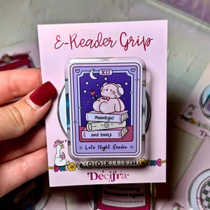 May include: A rectangular E-Reader Grip accessory on a pink display card. The centre features a purple tarot card illustration of a pink bear with headphones sitting on books, labelled Moonlight and books and Late Night Reader with the text Decifra.