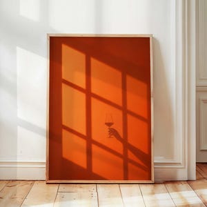 May include: A rectangular wall art print featuring a solid burnt orange background with a dark shadow cast by a window frame. The center of the print displays a silhouette of a hand holding a stemmed wine glass, creating a minimalist composition.