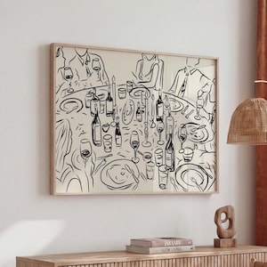 May include: Black and white line art print of a dinner party scene. The artwork depicts a round table set with plates, glasses, bottles, and candles. The print is framed in a light wood frame and hangs above a wooden cabinet.