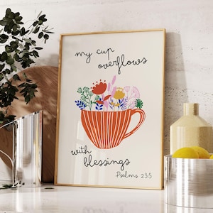 Op de afbeelding: Een ingelijste kunstprint toont een gestileerde oranje kop die overloopt van kleurrijke bloemen. De woorden "my cup overflows with blessings" staan boven en onder de kop, met "Psalms 23:5" onderaan. Het kunstwerk heeft een warme, uitnodigende uitstraling.