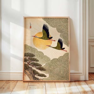 May include: Framed art print featuring two cranes in flight against a backdrop of a golden sun and swirling clouds. The artwork has a traditional Japanese aesthetic, with a detailed depiction of a tree on a cliff. The cranes are olive green with black wings and red beaks.