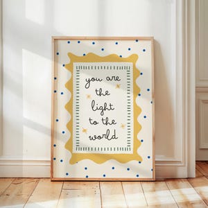 Puede incluir: Un cuadro enmarcado con fondo crema y lunares azules. La impresión presenta un borde amarillo ondulado alrededor del texto "you are the light to the world" en escritura negra. El marco es de color madera clara.