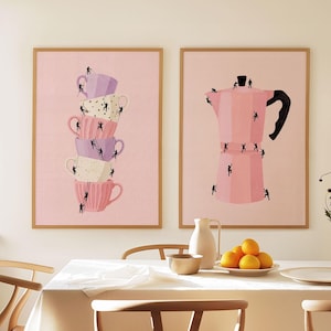 May include: Two framed art prints. One features a stack of pastel-colored teacups with tiny figures climbing ladders. The other depicts a pink coffee maker with similar climbing figures. Both are set against a soft pink background.
