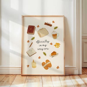 Könnte beinhalten: Ein gerahmter Druck mit Aquarellillustrationen von gemütlichen Gegenständen. Das Kunstwerk zeigt ein Buch, einen Pullover, Kaffee, ein Stück Kuchen, Hausschuhe und den Text "officially in my cozy era". Die Farbpalette ist warm, mit Brauntönen, Beige und Orange.
