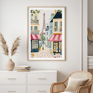 May include: Watercolor print of a Parisian street scene with the Eiffel Tower in the background. The artwork features buildings with pink awnings, flower boxes, and a cobblestone street. The print is framed in a light wood frame.