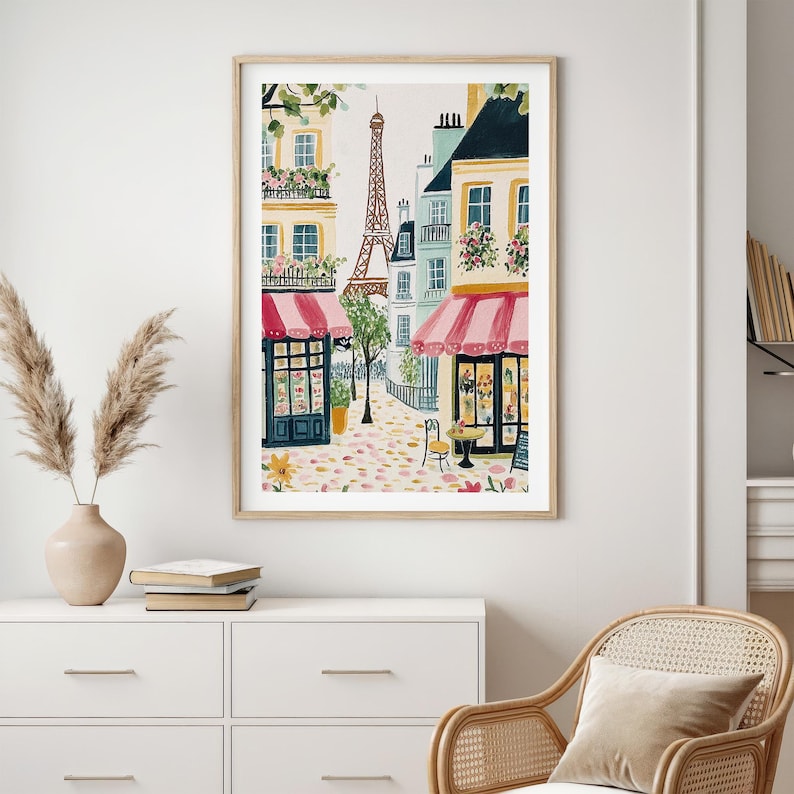 May include: Watercolor print of a Parisian street scene with the Eiffel Tower in the background. The artwork features buildings with pink awnings, flower boxes, and a cobblestone street. The print is framed in a light wood frame.