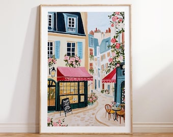 May include: A framed art print depicts a charming European street scene. Buildings with blue shutters, pink awnings, and blooming flowers line the cobblestone street. A cafe with outdoor seating and a chalkboard menu adds to the quaint atmosphere.