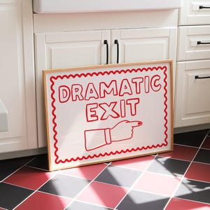 May include: A framed sign with the words "DRAMATIC EXIT" in red, along with a pointing hand graphic. The sign has a white background with a red scalloped border and a natural wood frame. The sign is leaning against white cabinets.
