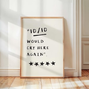 May include: A rectangular wall art print in a light wood frame featuring a cream background with black hand-drawn text that reads 10/10 WOULD CRY HERE AGAIN followed by five black hand-drawn stars arranged in a horizontal row at the bottom.