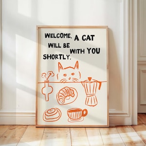 May include: A framed print with the text "WELCOME, A CAT WILL BE WITH YOU SHORTLY." The print features orange illustrations of a cat, coffee pot, croissant, and other breakfast items. The frame is a light wood colour.